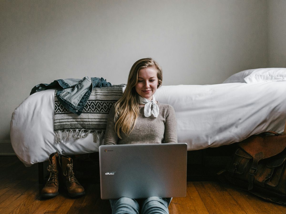 woman sitting beside a bed while using a laptop